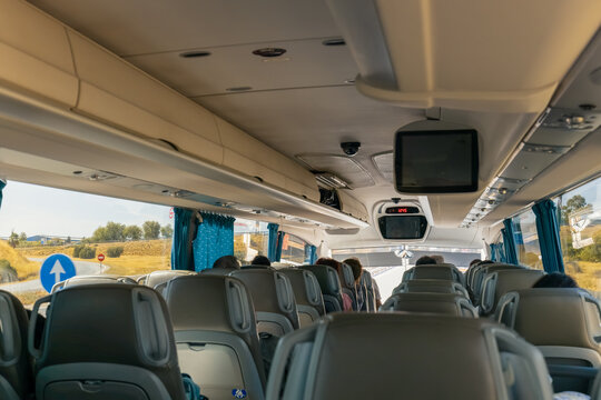 A modern passenger coach from a rear-row perspective. Several passengers are seated in high-back grey chairs as the bus travels along a sunny highway, visible through large side windows. 