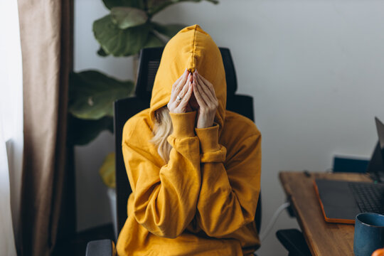 Depressed teenager hiding face in hoodie feeling loneliness and emotional isolation from social pressure in modern home interior