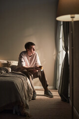 Vertical of Young adult Caucasian man sitting on bed holding mug, gazing thoughtfully out window with soft light illuminating face conveying emotion of contemplation and introspection