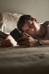 Obraz premium Vertical of Young Caucasian man lying on bed holding smartphone, gazing at screen with neutral expression, showing signs of emotional fatigue or contemplation in relaxed indoor setting