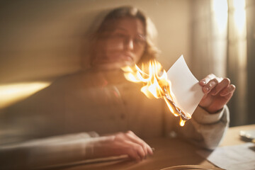 Caucasian young adult woman burning piece of paper while sitting at table, showing intense emotion through facial expression, blurred background emphasizing dramatic action