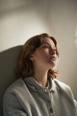 Vertical Portrait of young adult Caucasian woman leaning against wall, gazing upward with thoughtful expression conveying emotion in quiet indoor setting soft light highlighting facial features