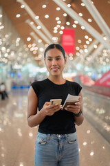 Young woman standing in airport terminal using phone and holding a passport with a boarding pass
