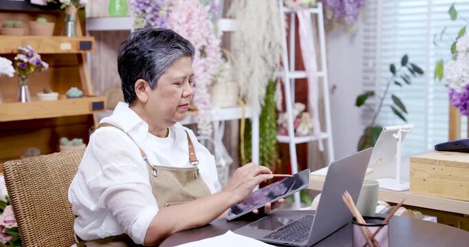 Asian elderly woman florist smiling while using tablet and laptop to manage flower shop ecommerce business seated in studio decorated with fresh and dried flowers multitasking with technology