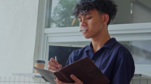 Portrait of young male student or creative professional writing in notebook while smoking