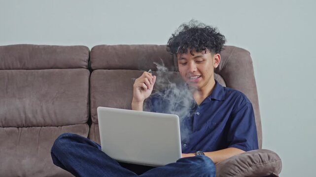 Casual lifestyle shot of curly-haired male sitting smiling while vaping with laptop on couch
