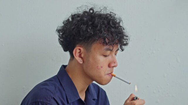 Close up of young Asian man lighting cigarette with lighter against white wall