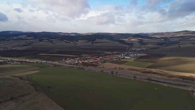 Žehra village in the Levoča District, Slovakia, appears as a tranquil rural settlement with red-roofed houses, patchwork fields, meadows and surrounding hills under an overcast sky from a drone view.