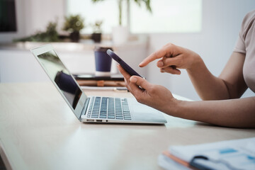 Obraz premium Close-up of person using smartphone beside open laptop on desk in bright modern office, remote work and digital communication concept.