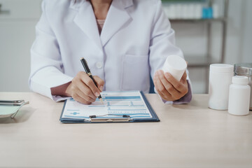 Fototapeta premium Doctor in white coat at desk writing on clipboard while holding medicine bottle, pharmacy containers on table, clinical office background