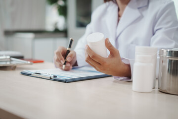 Healthcare professional in lab coat writing prescription while holding medicine bottle at clinic desk with other pill containers, close-up, copy space.