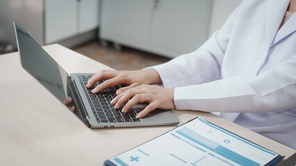 Doctor in white coat typing on laptop at desk with medical chart, modern clinic office, healthcare administration and digital patient records concept.