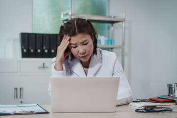 Stressed female doctor in white coat working on laptop at clinic desk, holding head with hand, paperwork and stethoscope nearby, healthcare burnout concept