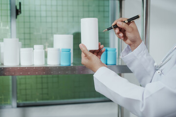 Pharmacist or lab technician in white coat holding a blank pill bottle and writing notes near a shelf of medicine containers in a clinical laboratory setting.