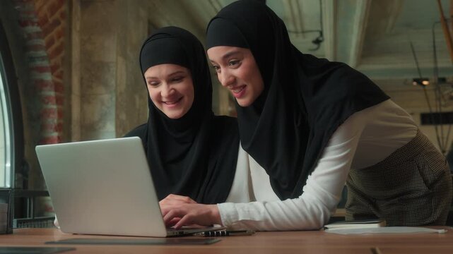Two happy smiling muslim islamic busines women wearing hijabs team girls businesswomen talking together looking in laptop computer screen teamwork company CEO teaching intern using software advice
