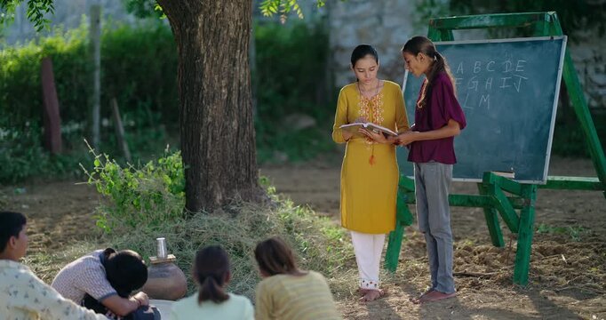 Indian young adult female hold book do study helping to read teen age girl class rural area place. Boy student group talk speak sitting on mat enjoy spend break time have fun joy open air ground