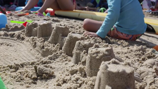 A young boy sits on the sandy beach and shapes a long row of sandcastles with colorful toys during a sunny day by the sea.
