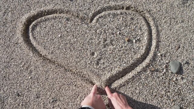 Couple drawing heart as a man and a woman use their fingers to sketch a large heart shape together on a gravel beach