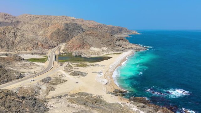 Aerial view of a coastal highway winding between rocky mountains and a sandy beach with turquoise water in Saudi Arabia.