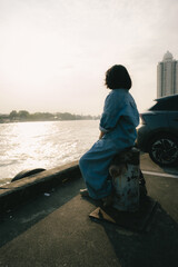 Fototapeta premium Portrait of asian woman in denim jumpsuit sitting on a river pier at the city skyline.