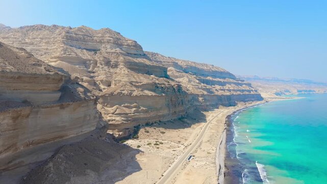 Aerial view of a long asphalt road stretching between massive desert cliffs and the turquoise waters of the Arabian Sea in Salalah, Oman.