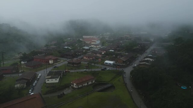 Atmospheric drone footage of Paranapiacaba historic district covered in dense morning fog. Traditional red-roofed houses line quiet streets in this preserved railway town near S&atilde;o Paulo, Brazil.