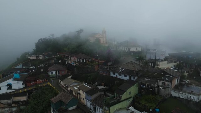 Atmospheric aerial footage of Paranapiacaba village emerging through dense fog in Santo Andr&eacute;, S&atilde;o Paulo, Brazil. Historic colonial architecture and church visible through misty mountain landscape.