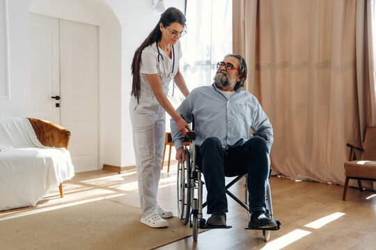 Female healthcare worker assists elderly man in wheelchair inside a well-lit room with wooden flooring and soft furnishings, providing support and care