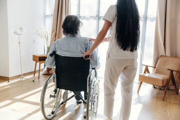 Fototapeta premium Caregiver assisting elderly man in wheelchair inside bright room with large windows, wooden floor, and comfortable seating area visible in the background