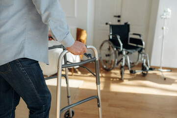 Fototapeta premium Senior man using walker in a bright room with wooden flooring, wheelchair visible in the background, emphasizing mobility assistance and home care environment