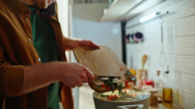 Cook hands adding chopped vegetables in soup talking smartphone home closeup