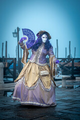 Obraz premium Venice, Italy - February 13, 2026: Woman in ornate purple and gold costume with mask and fan at Venice Carnival, foggy atmosphere with gondolas in the background