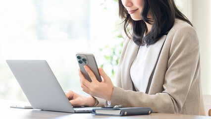 スマートフォンを見ながらノートパソコンを使うビジネスウーマン 業務効率や連携のイメージ, Businesswoman Using Smartphone and Laptop Multitasking Workflow Concept © Trickster*