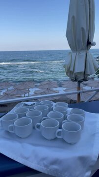 Rows of clean white coffee cups are set up on a table with a white cloth, overlooking a beach with many umbrellas and the blue ocean