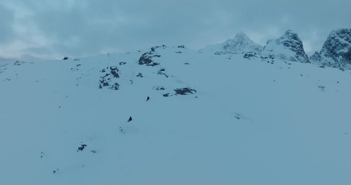 Stunning drone footage captures two skiers making their way across a pristine snow covered mountain valley in Norway. Dramatic jagged peaks rise majestically in the background under overcast winter