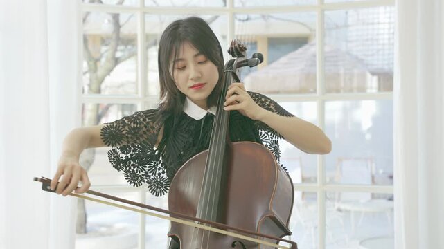 Beautiful young woman musician playing large cello with bow moving across strings with her choir. Female concentrating on playing cello while performing music with violinists in orchestra on stage