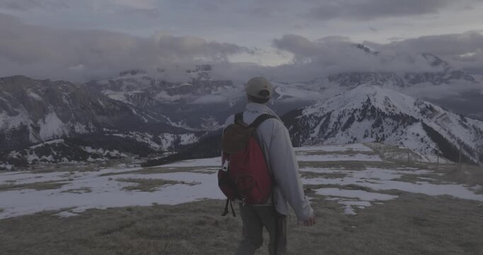 Lone hiker with red backpack walks through patchy snow covered alpine terrain during golden hour. Dramatic snow capped mountain peaks stretch across horizon under moody cloudy sky. Peaceful winter