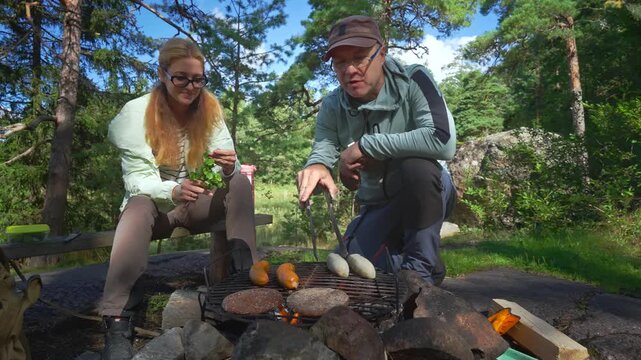 Young couple grilling sausages and burgers over campfire in sunny forest. Authentic outdoor culinary experience, sustainable living and mental wellness in a Finnish national park.