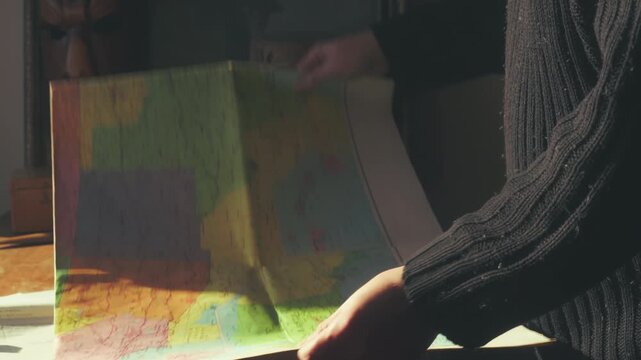 Medium shot of a person unfolding a large, colorful vintage travel map of the United States on a warm, sunlit table. Evokes trip planning vibes.