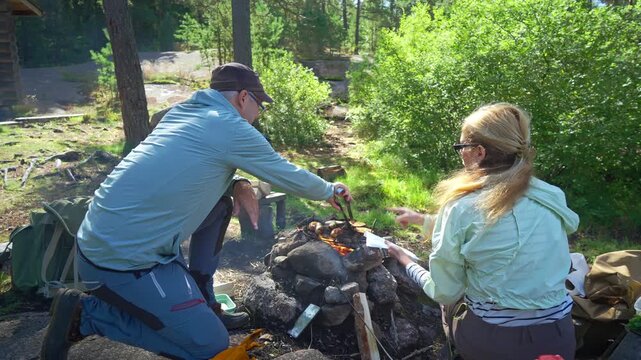 Young couple grilling sausages and burgers over campfire in sunny forest. Authentic outdoor culinary experience, sustainable living and mental wellness in a Finnish national park.