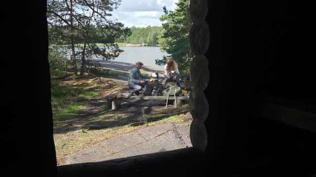 Active couple with backpacks arriving at wooden shelter in Finnish national park. Responsible eco-tourism, wilderness infrastructure and authentic Nordic hiking lifestyle in sunny forest.