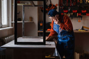 Female carpenter in overalls talking on phone and taking orders in a small craft workshop.