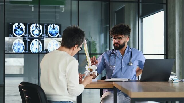 Male doctor discusses spinal structure with female patient using a model in a contemporary medical office featuring X-ray images on the wall