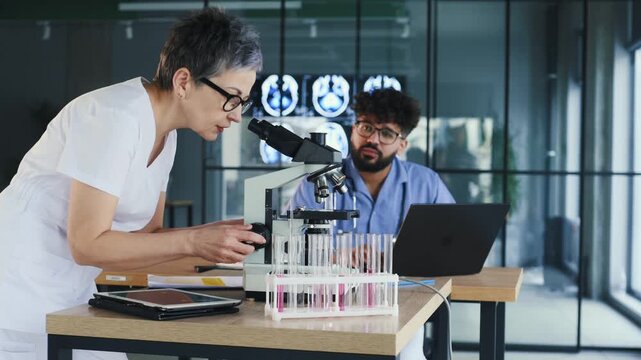 Female scientist in lab coat analyzes samples with microscope, while male coworker uses laptop in a contemporary laboratory environment