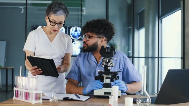 Female healthcare worker in white uniform talks to male colleague in blue scrubs at a laboratory table with a microscope and test tubes in a medical facility