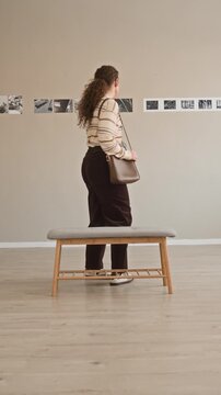 Vertical full stab shot of young Caucasian woman listening to audio guide in earphones taking seat on bench, observing black and white fine art photography exhibition in modern gallery