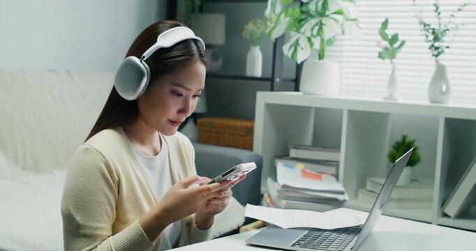Modern Asian person using a smartphone and laptop for finance. She is checking a bank statement connection and managing her personal economy indoors.