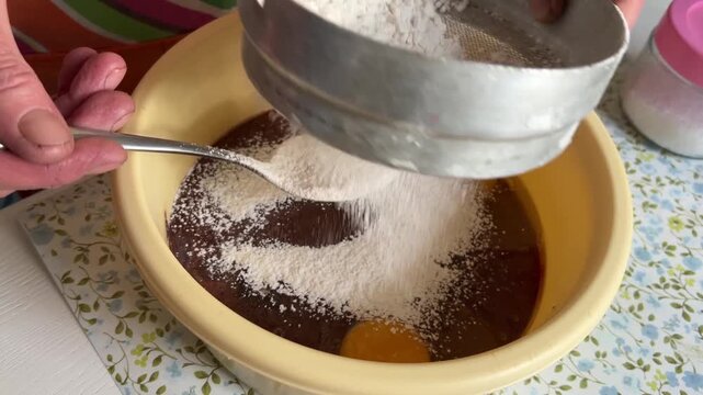 Horizontal slightly top view video: flour is sifted through a sieve and gradually added with a tablespoon to the mixture, cooking process