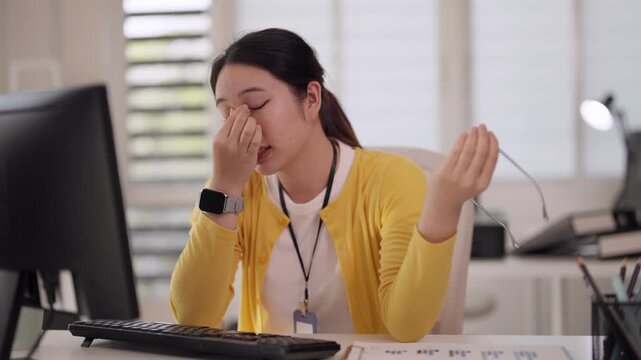 Asian adult woman experiences significant work stress and burnout at office desk holding eyeglasses contemplating job challenges