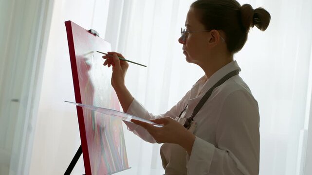 Skilled female painter creating expressive artwork on easel in bright studio. Artist depicting colorful portrait during creative process inside professional workshop.